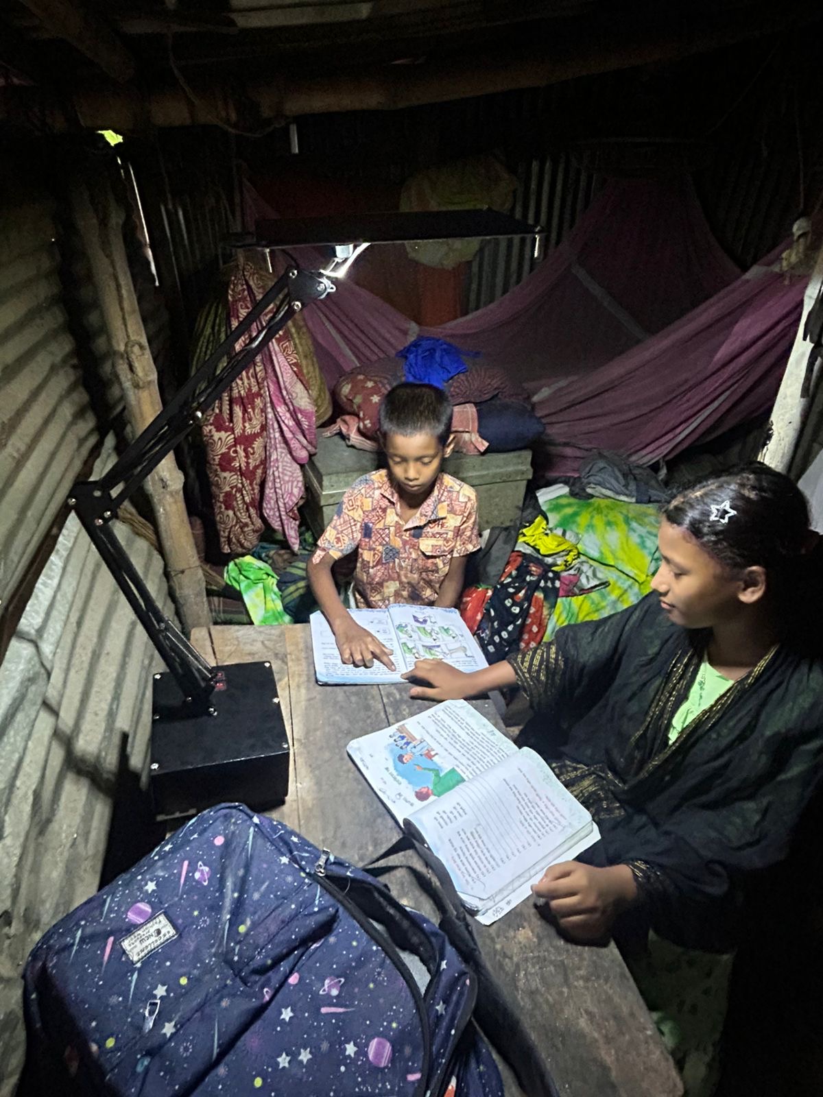 Two siblings studying at night under a small solar-powered LED lamp; notebooks open.